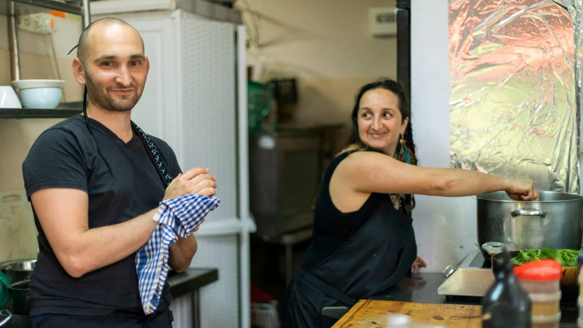 קאמה עומד במטבח של בית הקפה ומנגב ידיים במגבת, ואפרת מאחוריו מבשלת בסיר גדול
